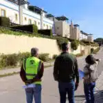 Três pessoas caminham em rua residencial ensolarada.