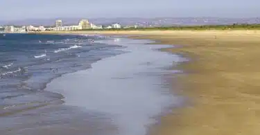 Praia extensa com ondas suaves e cidade ao fundo.